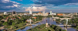 Aerial view of the downtown Wichita, Kansas skyline against a blue sky, with the Arkansas River in the foreground