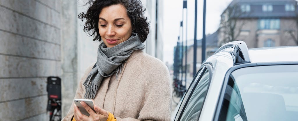 A woman standing next to her car smiles while looking at her phone because she received her car insurance refund.