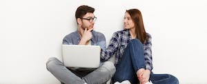 Young couple leaning against a white backdrop with a laptop while discussing whether they should refinance their mortgage